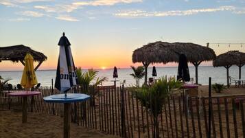 A beach with thatched umbrellas and tables, a wooden fence, and a sunset in the background.