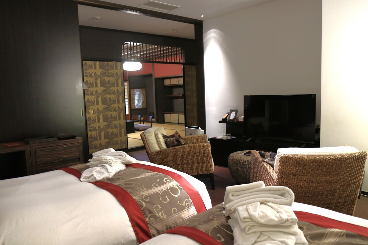 A hotel room with a bed, two chairs, a TV, and a desk with a lamp.
