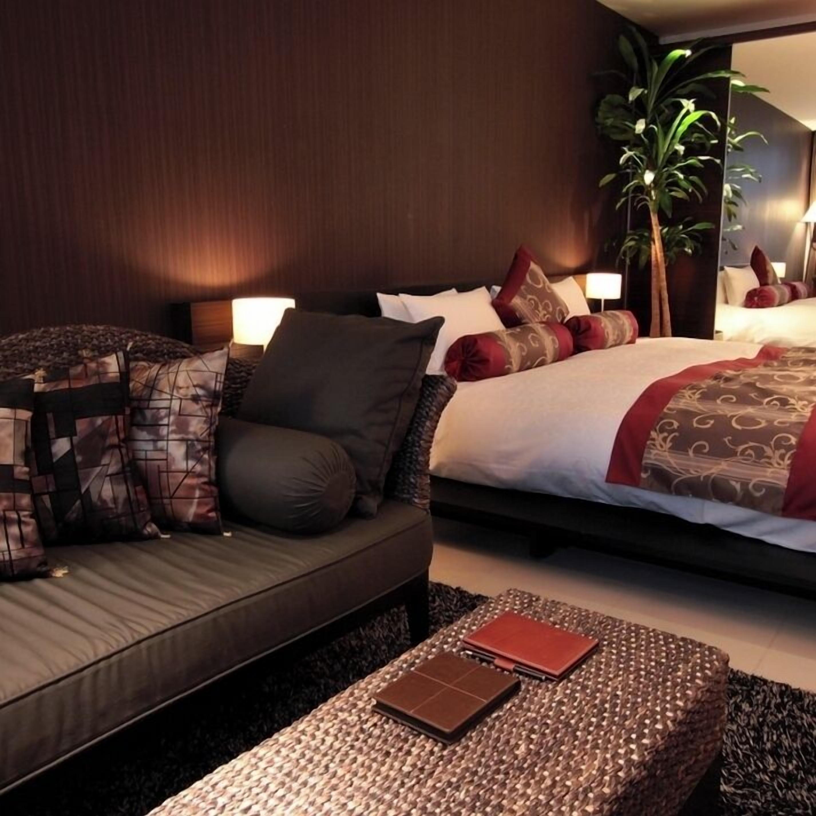 Et moderne hotelværelse med sengegavl i mørkt træ, en stor seng med mønstret sengetøj, en mørk sofa med mange puder, et lille bord med en bog og en potteplante.