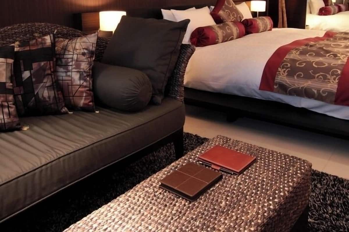 A modern hotel room with a dark wood headboard, a large bed with patterned bedding, a dark sofa with multiple cushions, a small table with a book, and a potted plant.