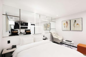 Quarto de hotel moderno com uma cama grande, área de estar com poltrona e mesa, escrivaninha com computador e obras de arte nas paredes.