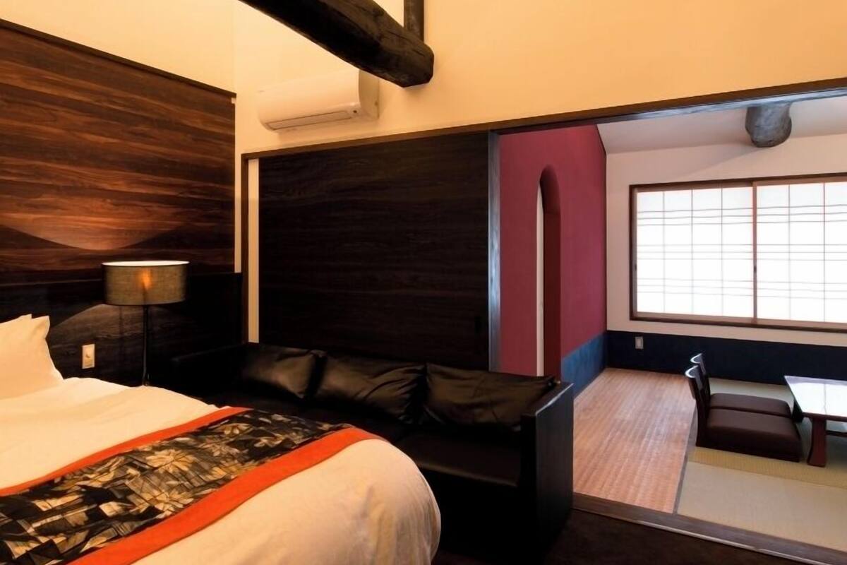 A hotel room with a large bed, a desk, a chair, a sofa, and a view of a traditional Japanese room.