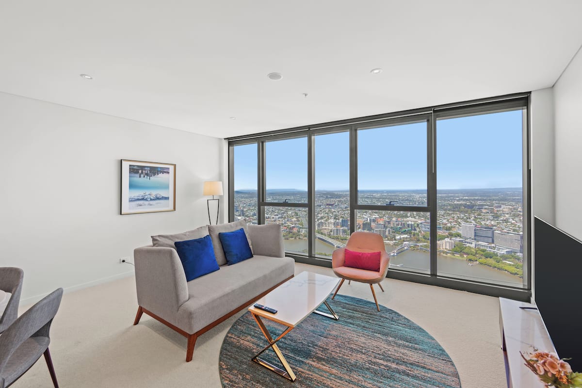 A modern living room with a sofa, armchair, and coffee table, large windows with a city view, and a framed picture on the wall.
