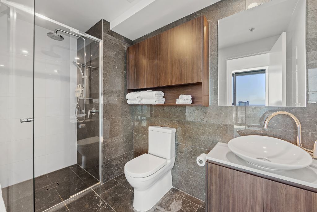 Une salle de bain moderne avec une douche à parois de verre, des toilettes blanches, un sol carrelé en marbre, un meuble-lavabo en bois doté d’un grand évier et un miroir au-dessus du meuble-lavabo.