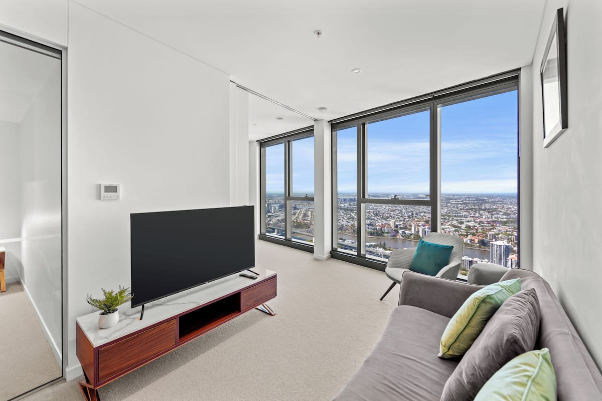 A modern living room with a large flat-screen TV, a grey sofa with cushions, and a city view through large windows.