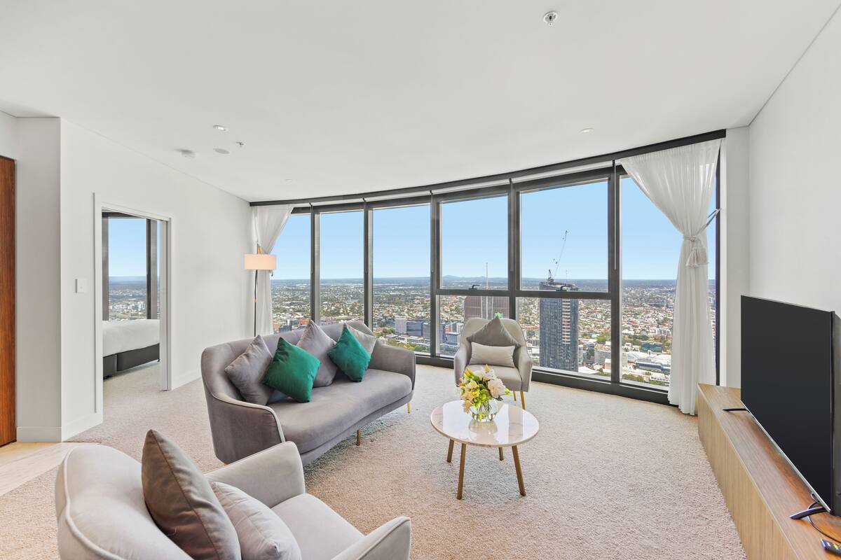 A spacious living room with a large window offering a city view, a grey sofa with green cushions, a round coffee table with flowers, and a flat-screen TV on a wooden stand.