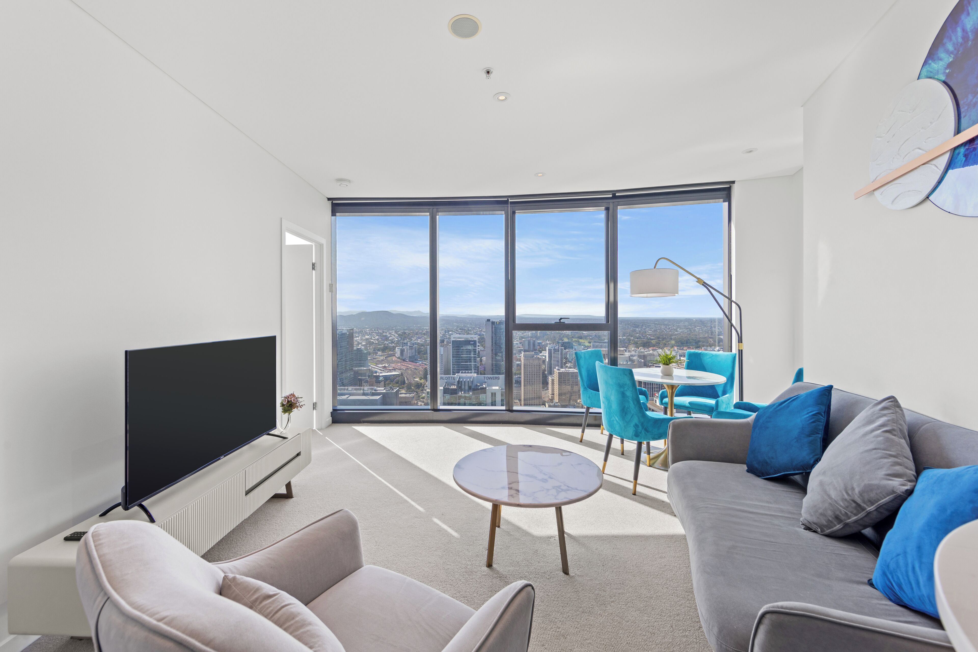 A modern living room with a large TV, comfortable seating, a small table, and a city view through large windows.