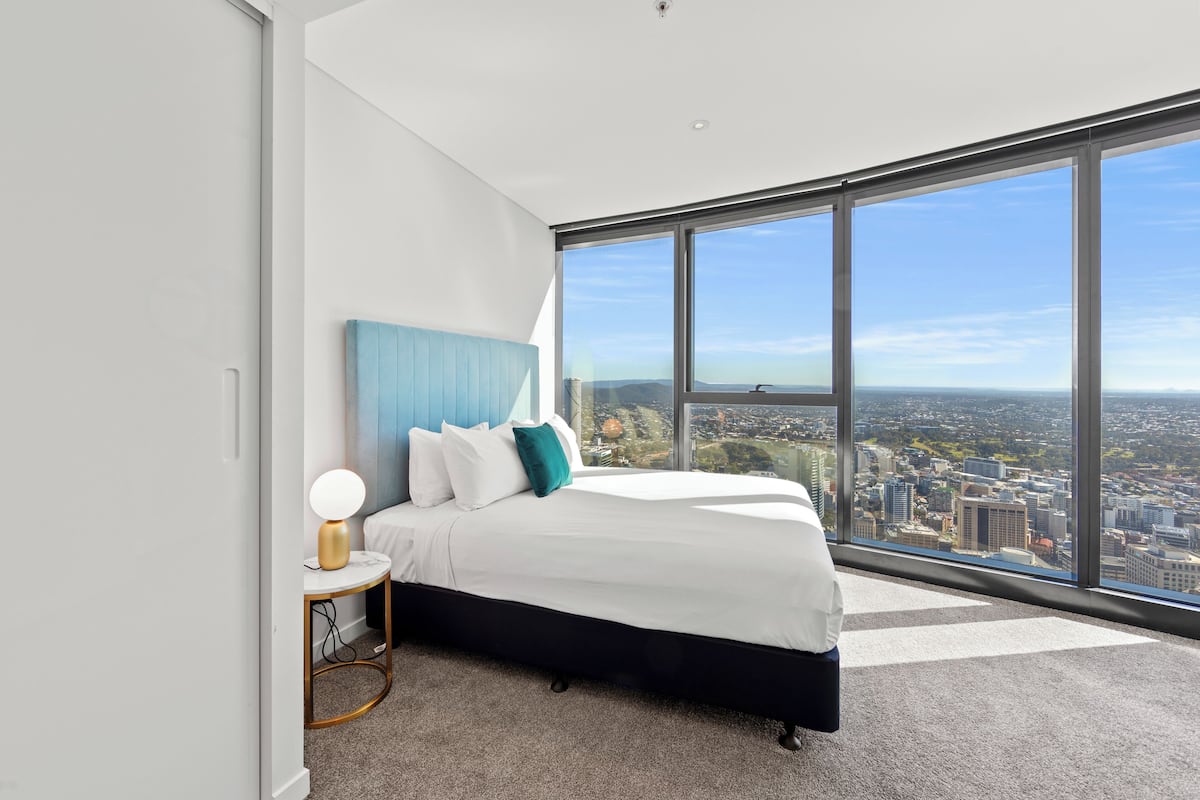 A modern bedroom with a large bed, a nightstand, and a view of a cityscape through floor-to-ceiling windows.