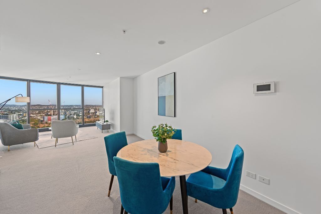 Una habitación moderna con una mesa redonda de mármol, sillas azules, una planta en maceta y vistas a la ciudad a través de amplios ventanales.