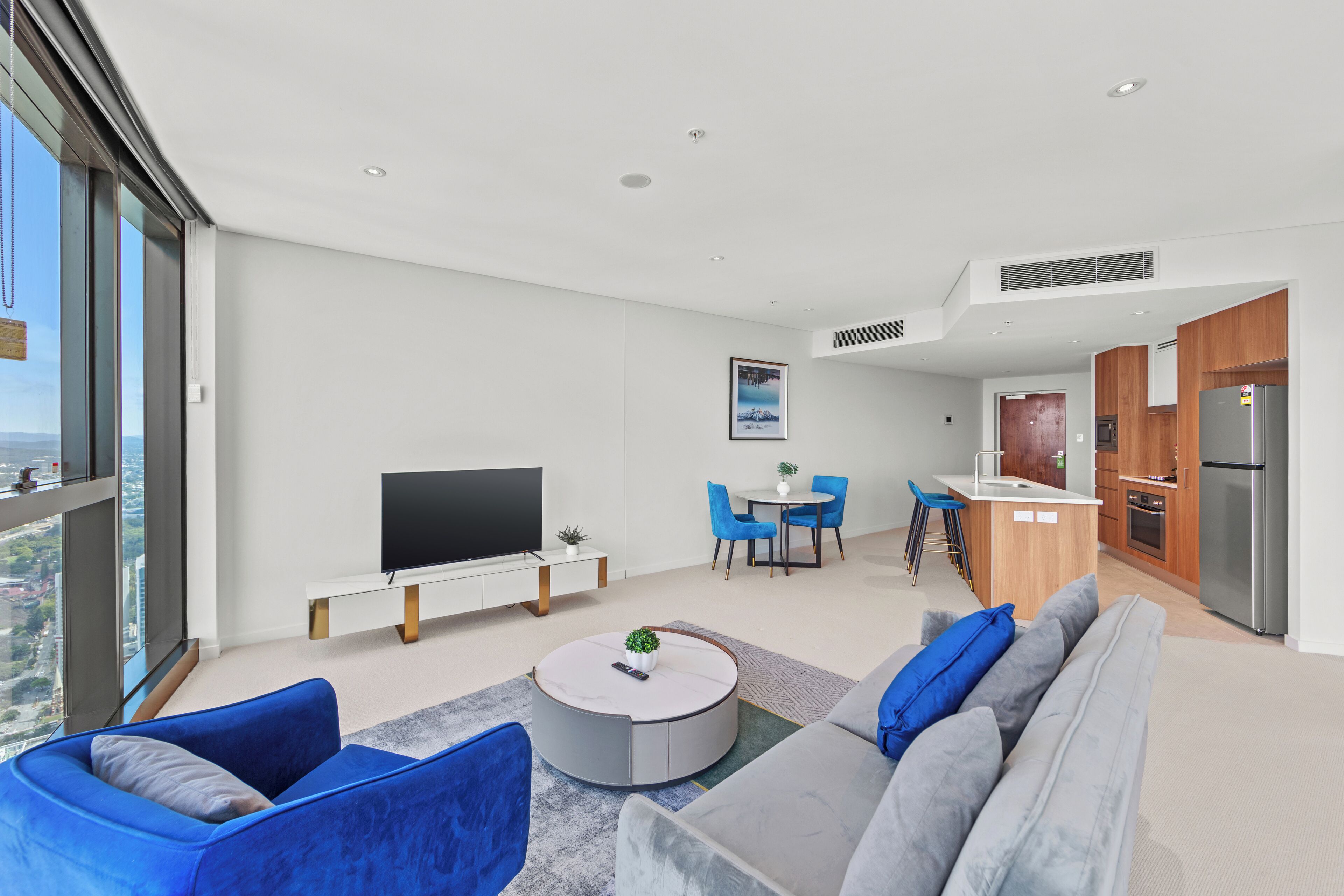 A modern living room with a flat-screen TV, a round coffee table, and a sofa. There is a dining area with blue chairs and a small kitchen with a refrigerator and a sink.