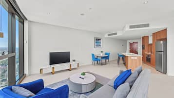 A modern living room with a flat-screen TV, a round coffee table, and a sofa. There is a dining area with blue chairs and a small kitchen with a refrigerator and a sink.