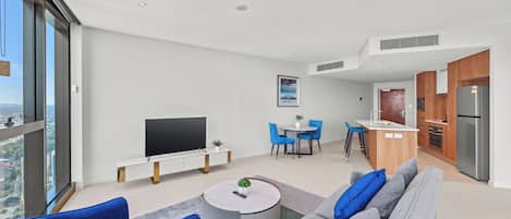A modern living room with a flat-screen TV, a round coffee table, and a sofa. There is a dining area with blue chairs and a small kitchen with a refrigerator and a sink.