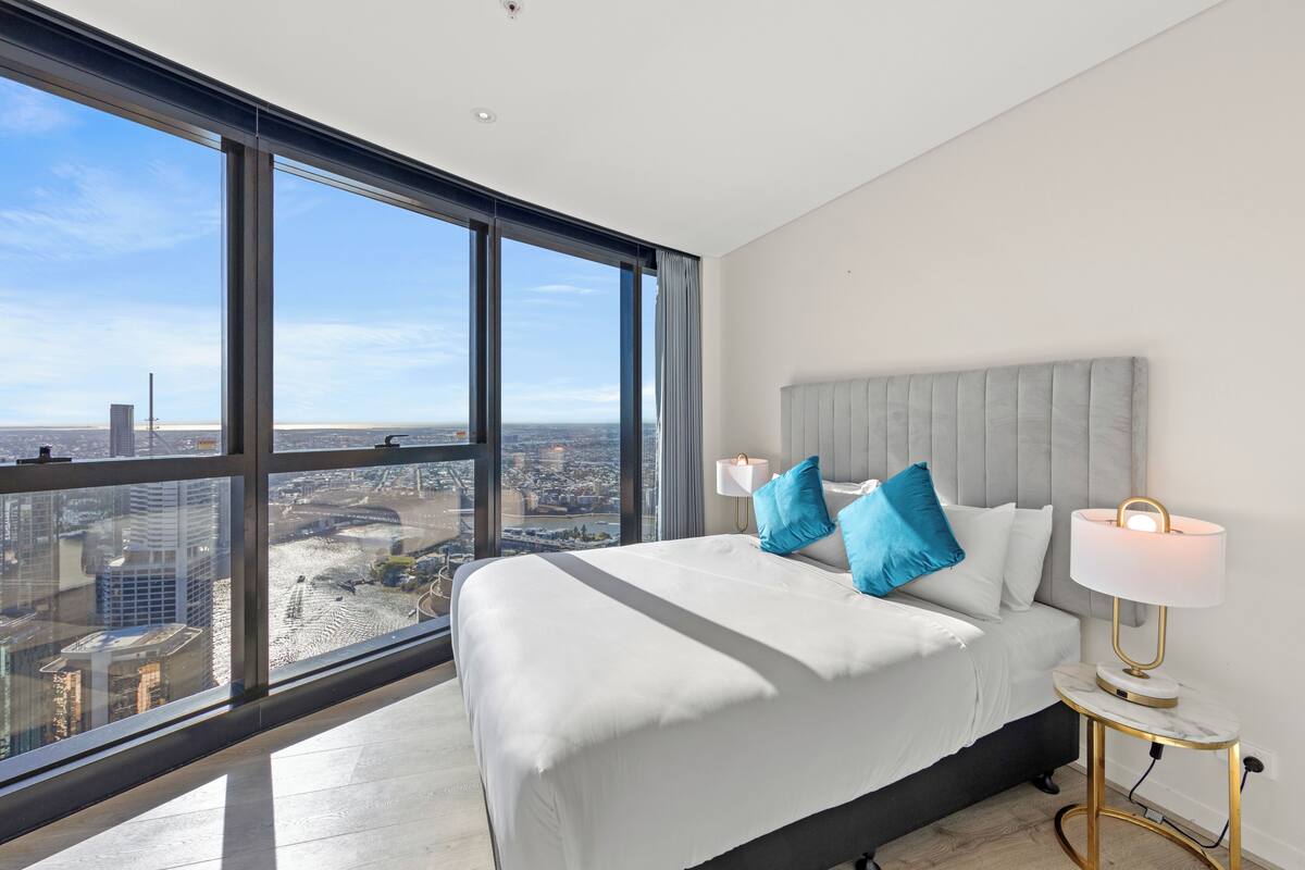 A modern bedroom with a large window, a bed with white bedding and blue pillows, and a bedside table with a lamp.