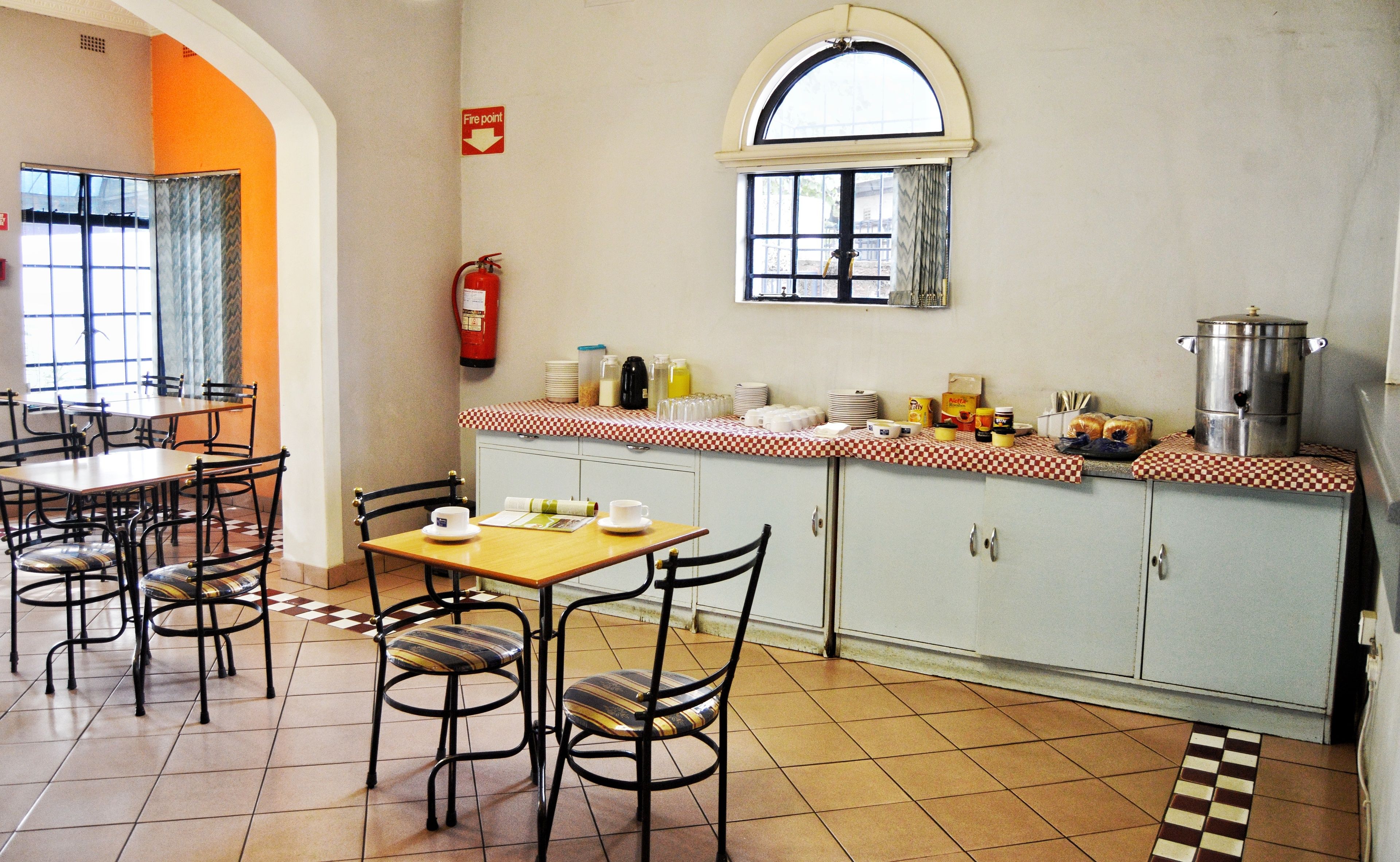 Un comedor con mesas y sillas, un extintor, una ventana y una encimera de cocina con diversos objetos.