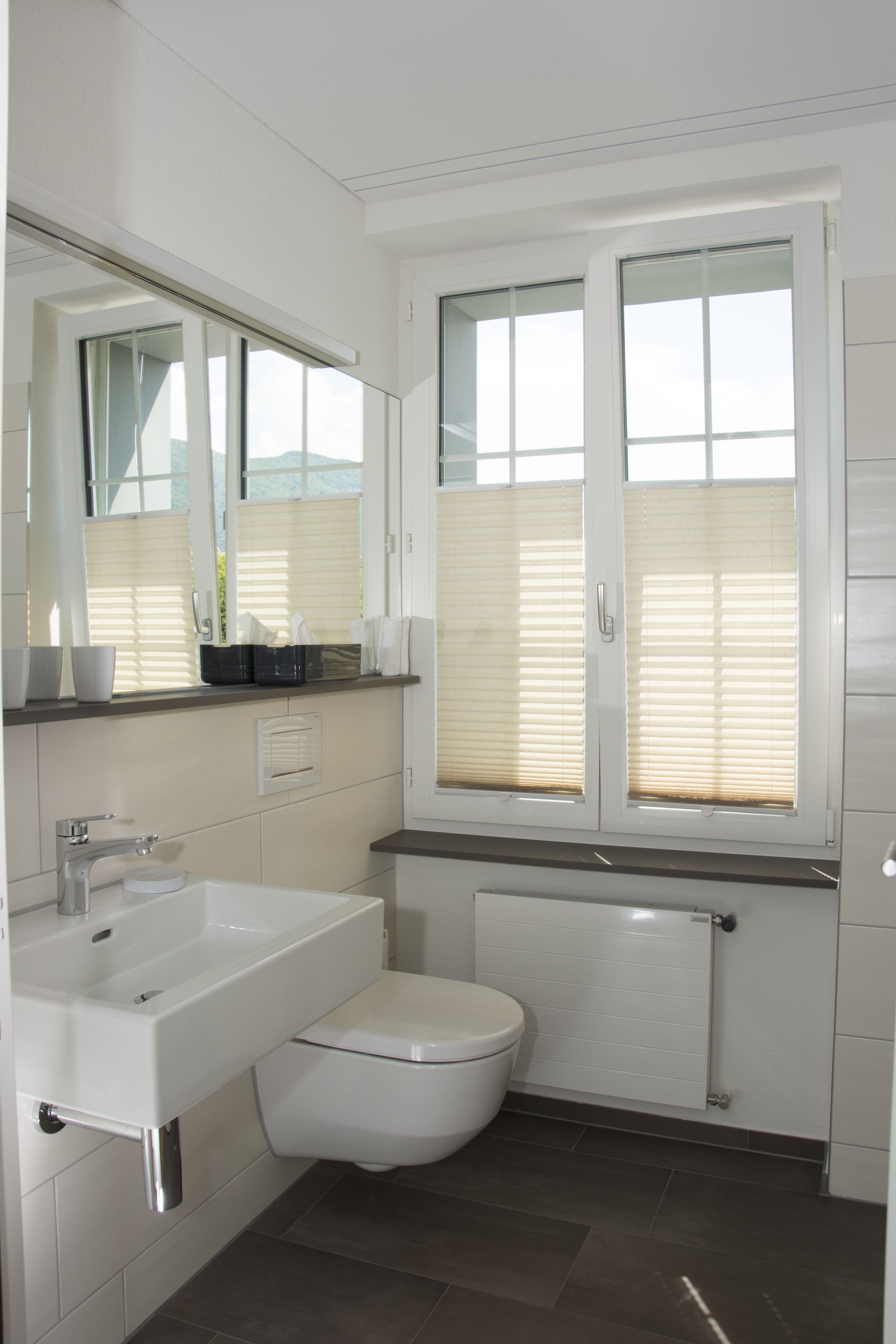 A modern bathroom with a white sink, toilet, and a large mirror. There is a window with blinds and a radiator below it.