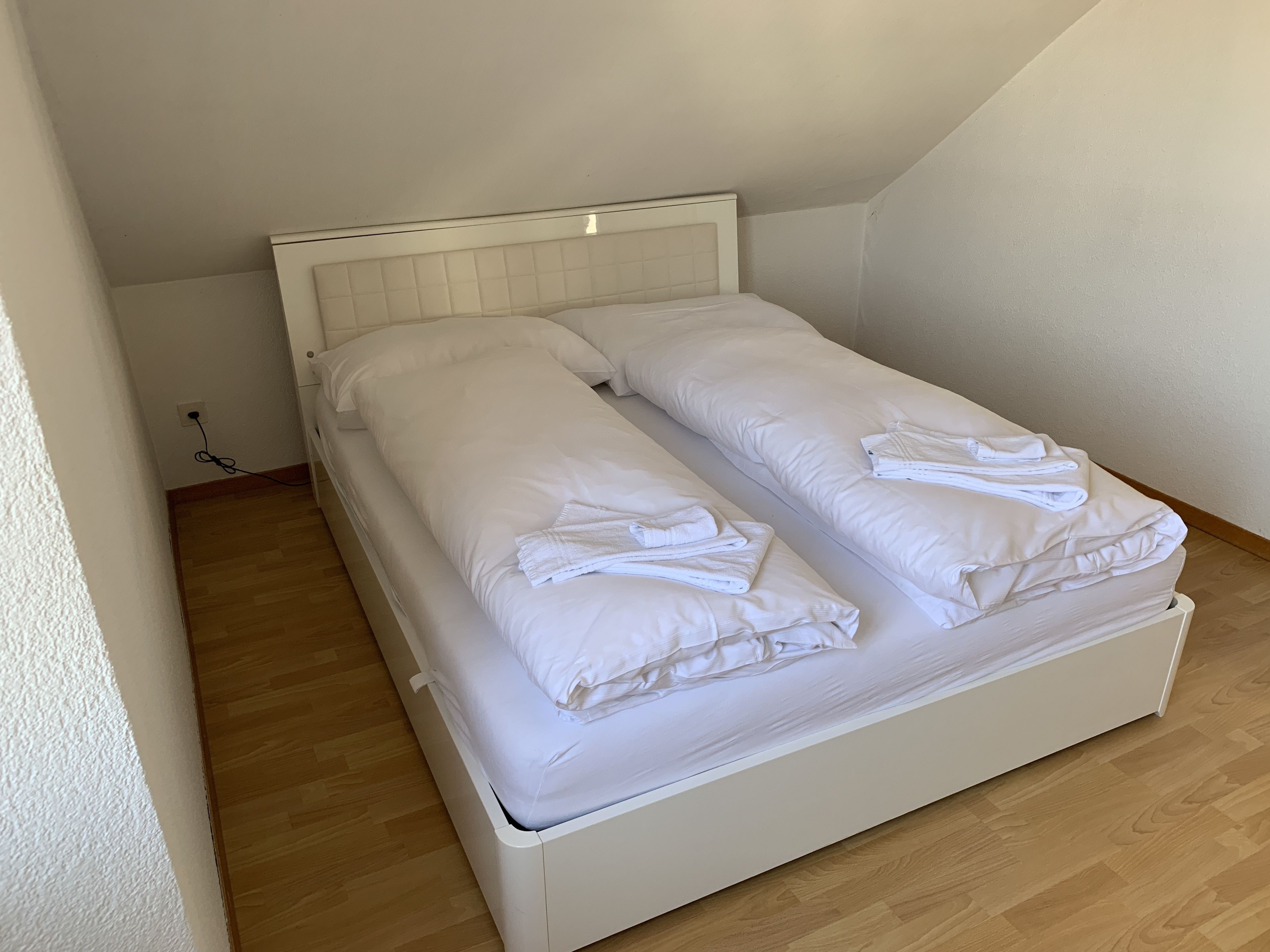 A single bed with white bedding and towels, a wooden floor, and a white headboard.