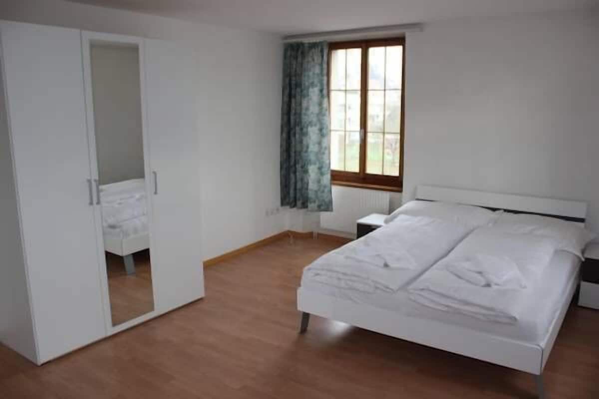 Quarto com cama, guarda-roupa, janela com cortinas e mesa de cabeceira.