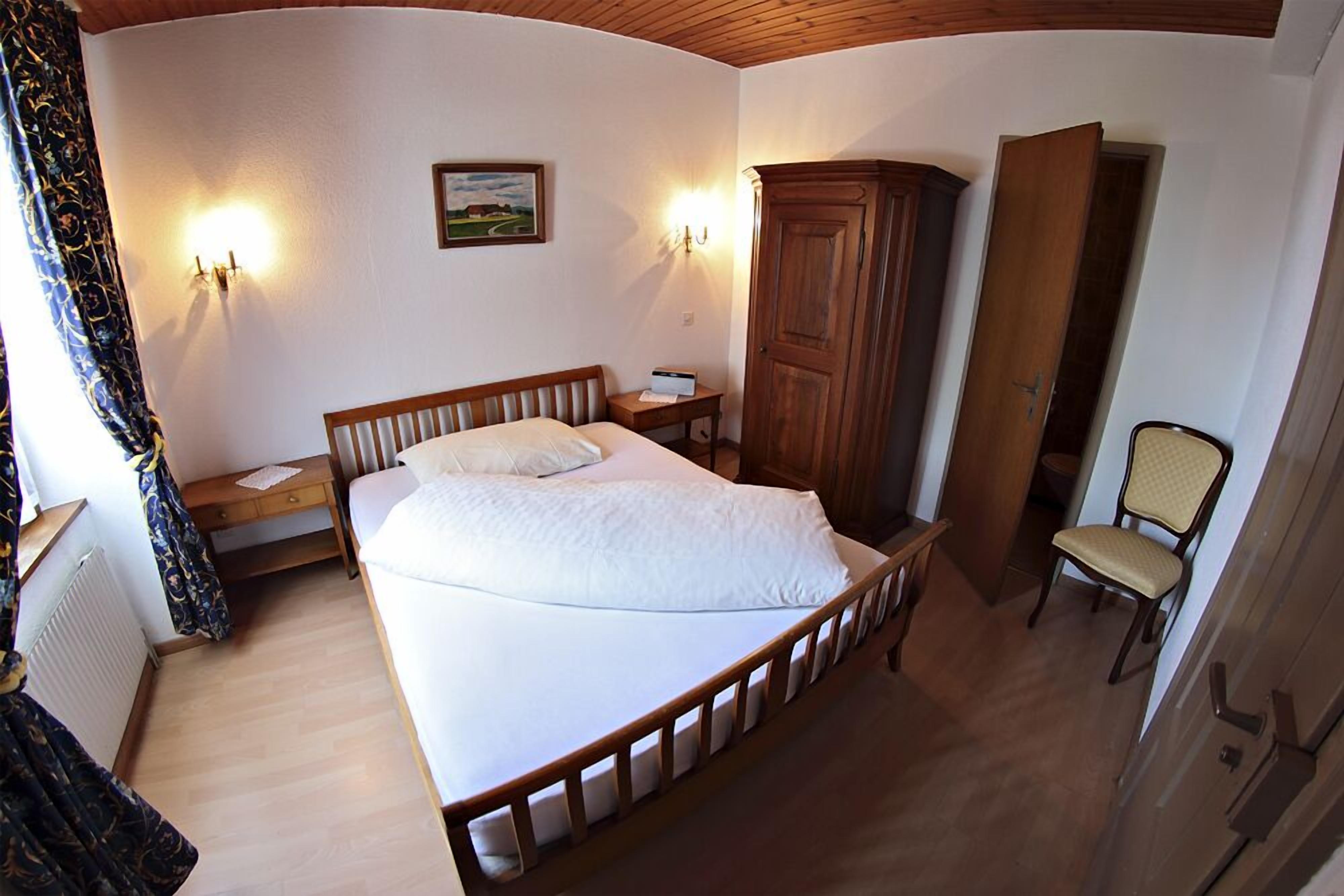 A bedroom with a bed, a wooden wardrobe, a chair, and two wall-mounted lamps.