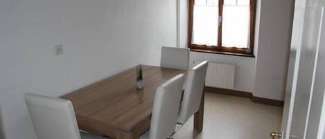 A dining area with a wooden table and four chairs, a window with blinds, and a radiator.