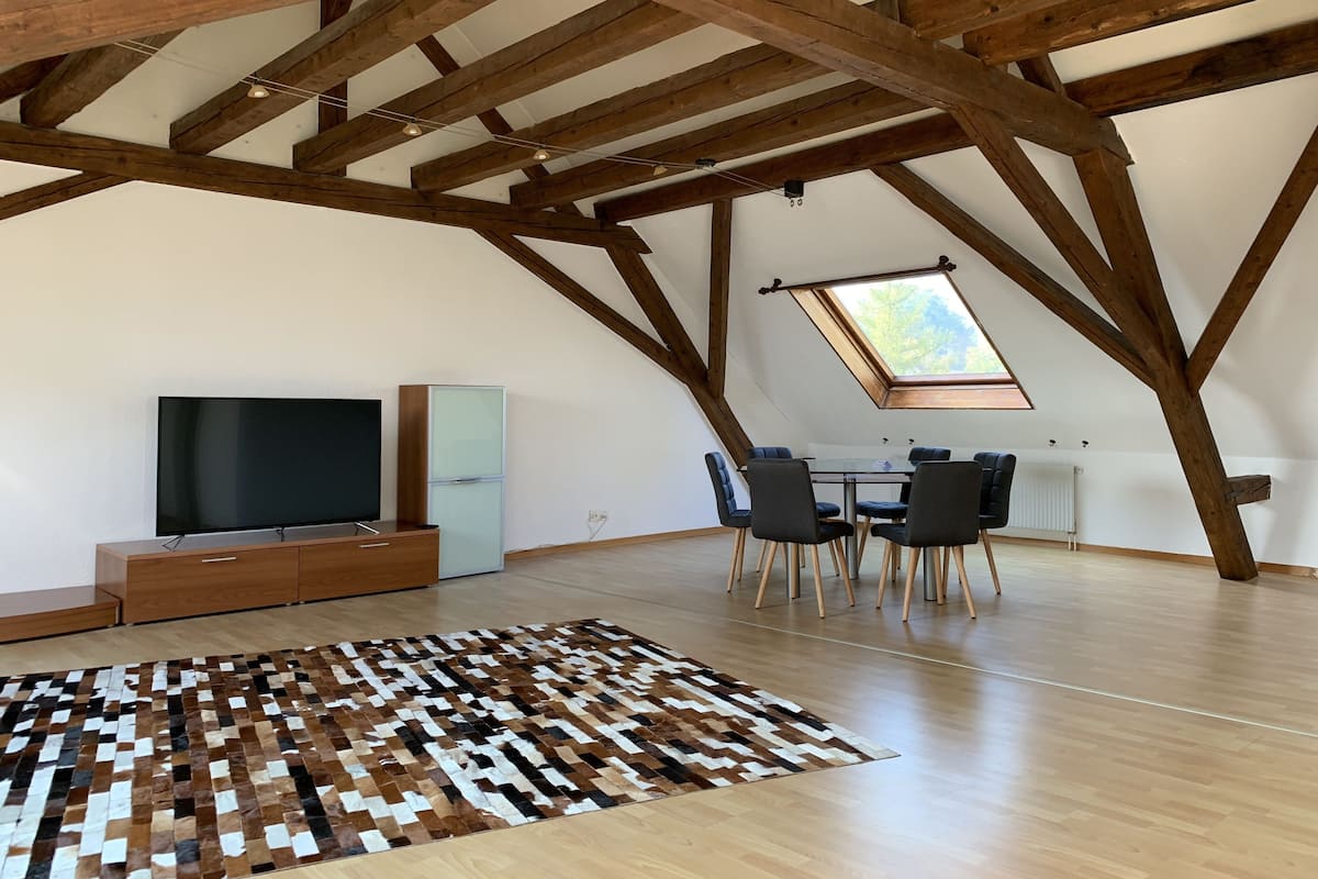 Um quarto amplo no sótão, com vigas de madeira aparentes, uma televisão grande, uma mesa de jantar com cadeiras e um tapete estampado.