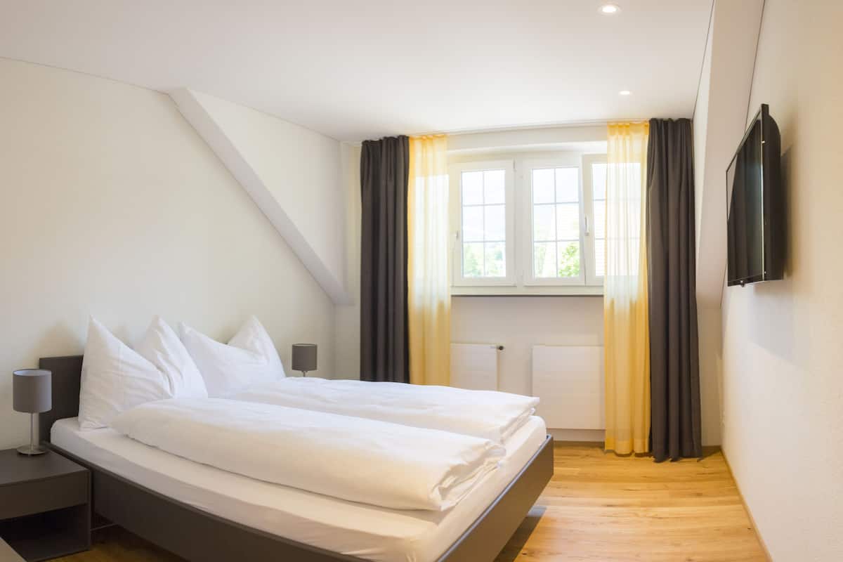 Quarto de hotel moderno com cama, mesas de cabeceira, janela com cortinas e televisão de tela plana na parede.