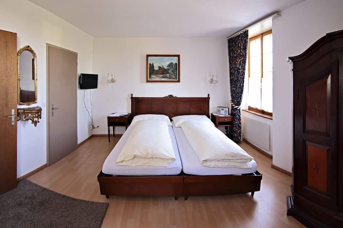 Uma cama de casal com cabeceira de madeira, uma mesa de cabeceira, uma televisão na parede e uma janela com cortinas.