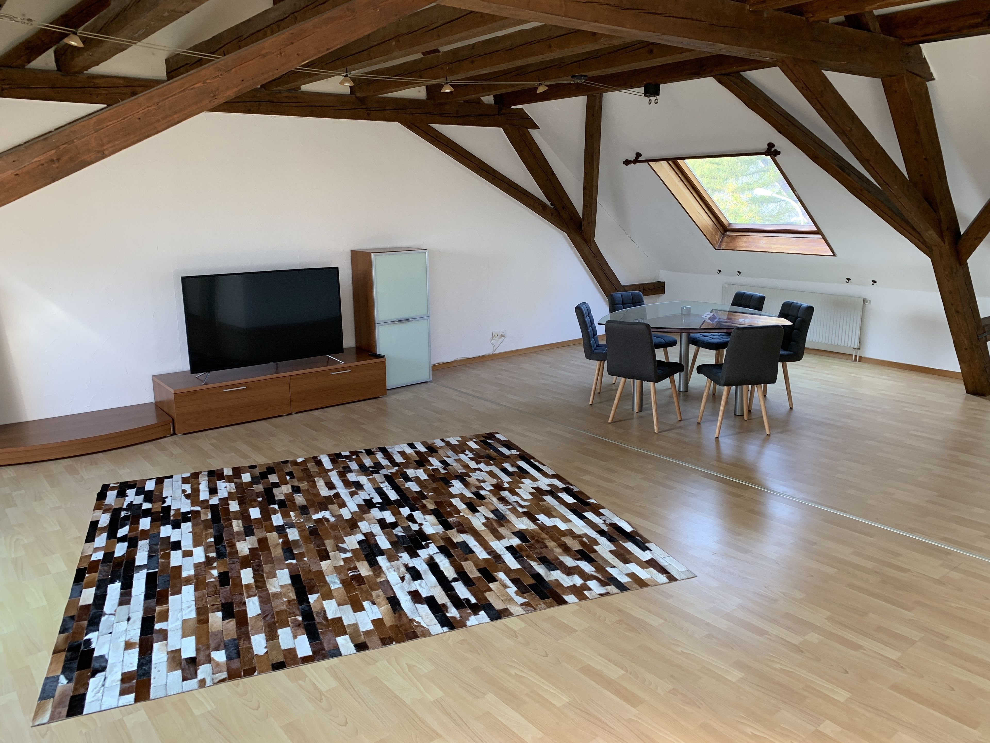 A spacious loft with a large wooden ceiling, a flat-screen TV, a glass dining table with chairs, and a patterned rug.