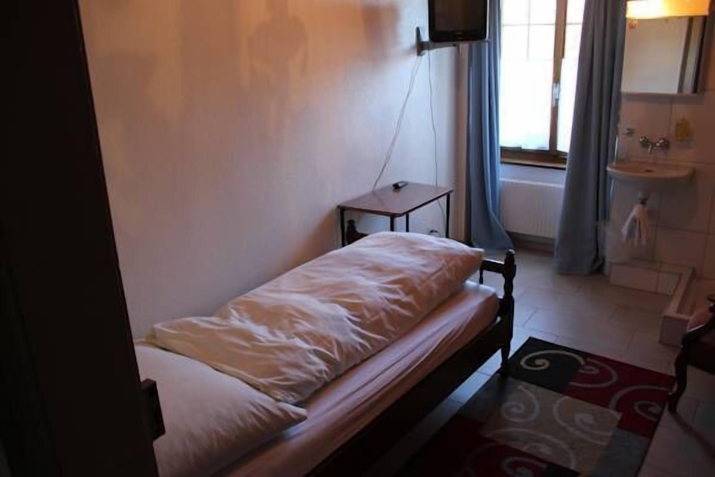 Une petite chambre avec un lit simple, un lit en bois, une petite table de chevet, un téléviseur fixé au mur, une fenêtre avec des rideaux et une salle de bain avec un lavabo et un miroir.