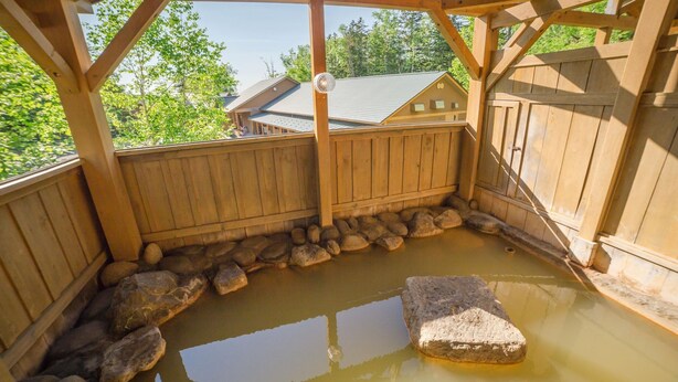 A wooden structure with a stone-lined pond, wooden beams, and a view of a house in the background. - Biei Guest House Sirogane no Yu - Hostel (Biei)