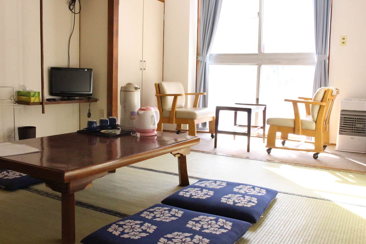 Perinteinen japanilaistyylinen huone, jossa on tatamimatto lattiana, matala puupöytä, kaksi kukka-aihekuvioitua tatamimattoa, hyllyllä oleva televisio ja ikkuna, jossa on verhot.