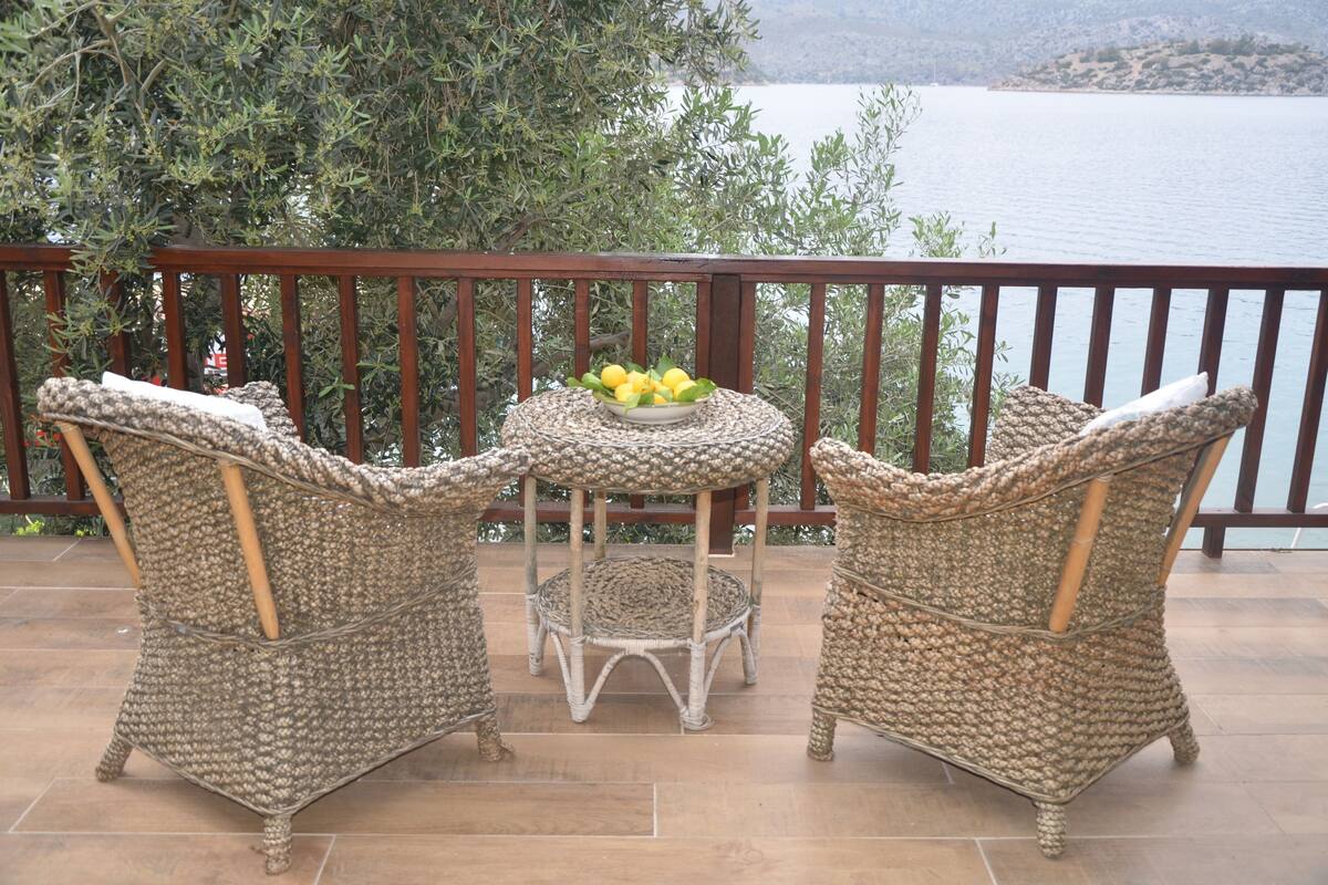Een set rieten tafels en stoelen op een balkon met uitzicht op een meer en bomen.