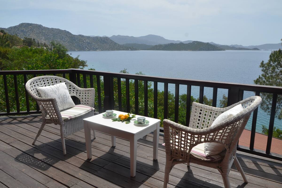 Een terras met rieten stoelen en een tafel gedekt voor twee, met uitzicht op een meer en bergen op de achtergrond.