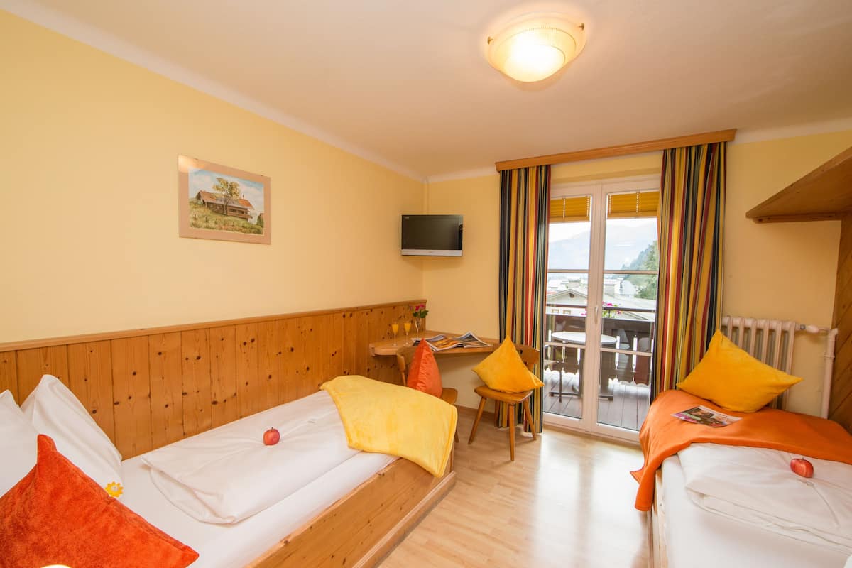 A hotel room with two single beds, a wooden headboard, a television, a small table with a chair, and a window with curtains.