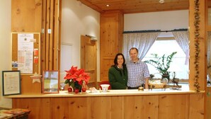 A wooden reception desk with a bulletin board, a potted plant, and a window with curtains. - Pension Hubertus (Zell am See)