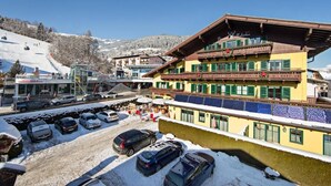 A hotel with a ski lift in the background, a parking area with cars, and a snowy mountain landscape. - Pension Hubertus (Zell am See)