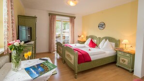 A hotel room with a bed, bedside tables, a TV, a window with curtains, a vase with flowers, and a table with a book and glasses. - Pension Hubertus (Zell am See)