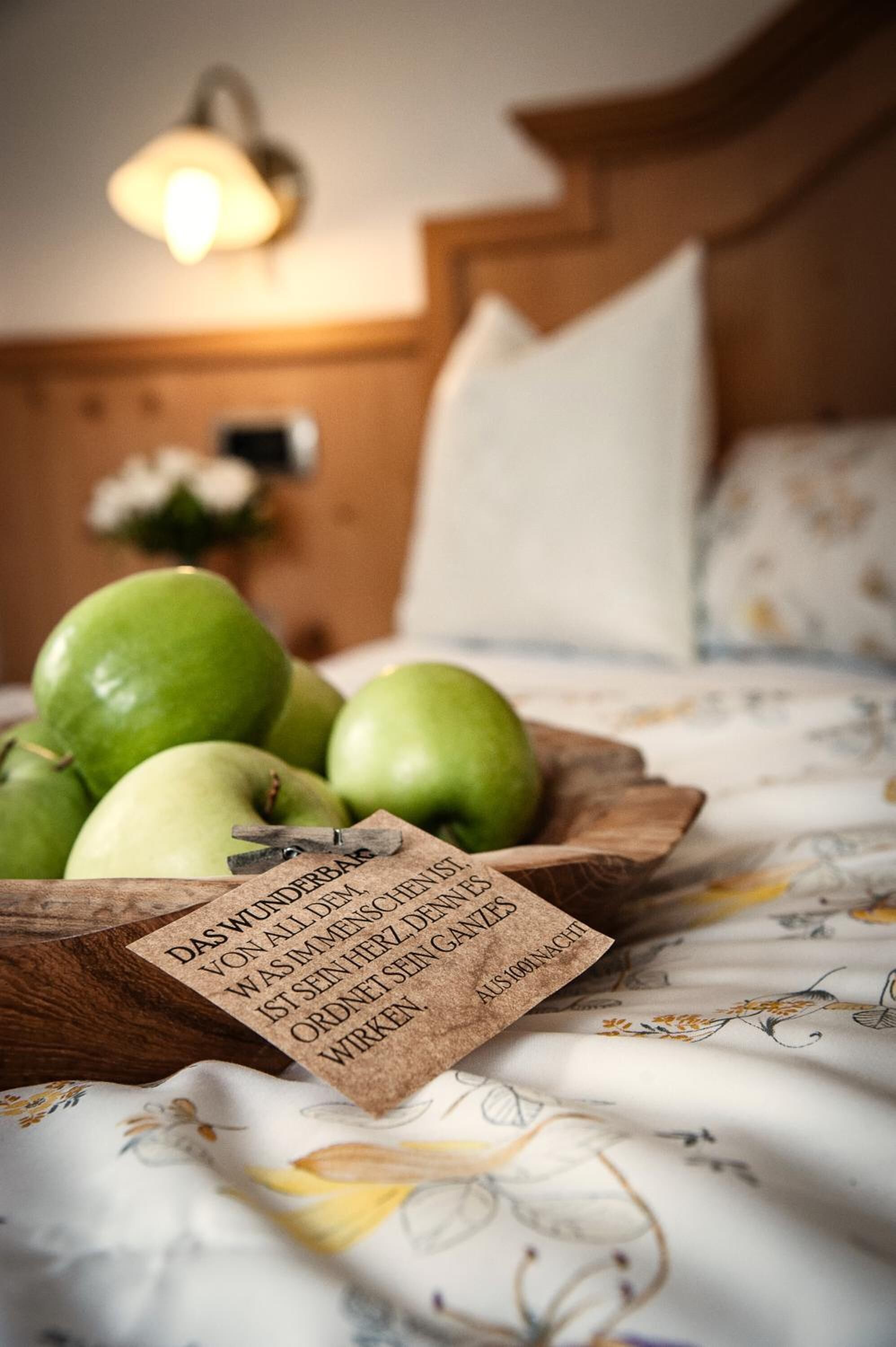 A wooden bowl with green apples on a bed with a floral pattern.