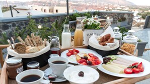 Un plateau de petit-déjeuner comprenant du pain, des viennoiseries, des fruits, du fromage et des boissons, servi sur une terrasse offrant une vue sur la ville.