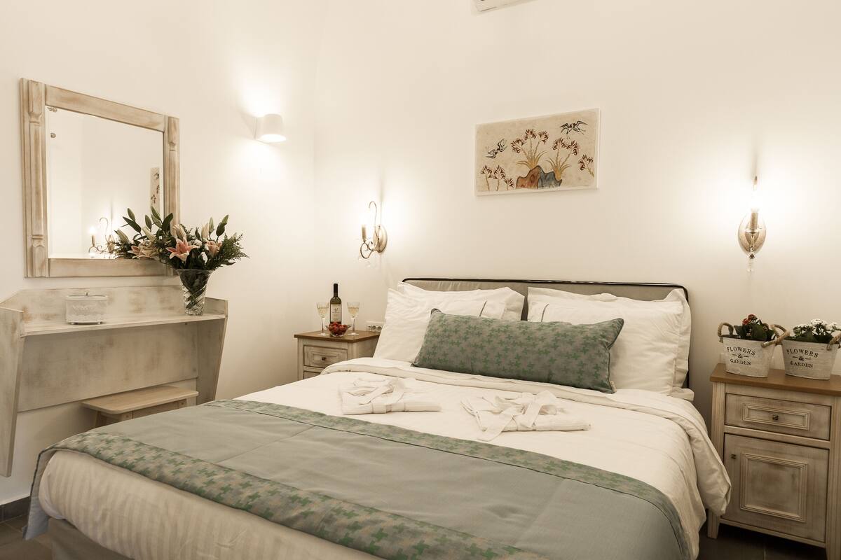 Ein ordentlich gemachtes Bett mit einer grün-weißen Tagesdecke, ein Nachttisch mit Blumen in einer Vase und ein an der Wand befestigter Spiegel.