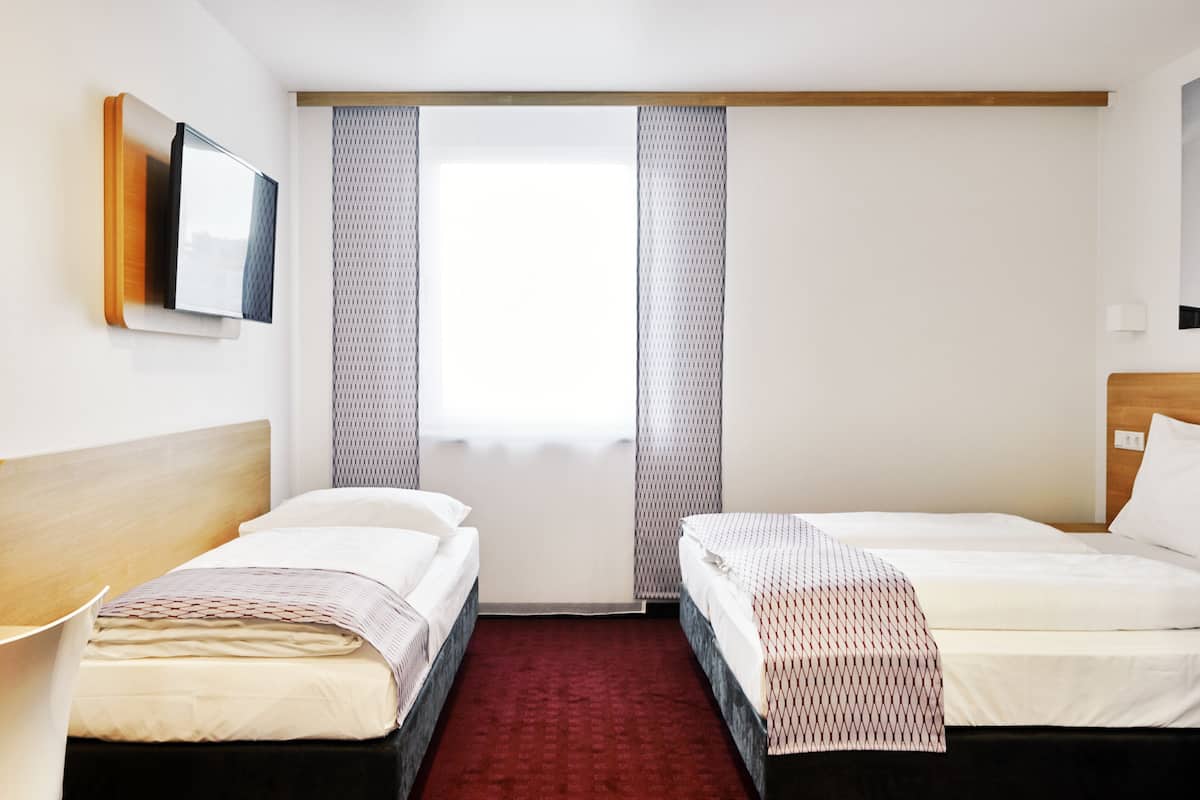 Une chambre d’hôtel avec deux lits simples, une tête de lit en bois, un téléviseur à écran plat fixé au mur, une fenêtre avec des rideaux à motifs et une moquette rouge.