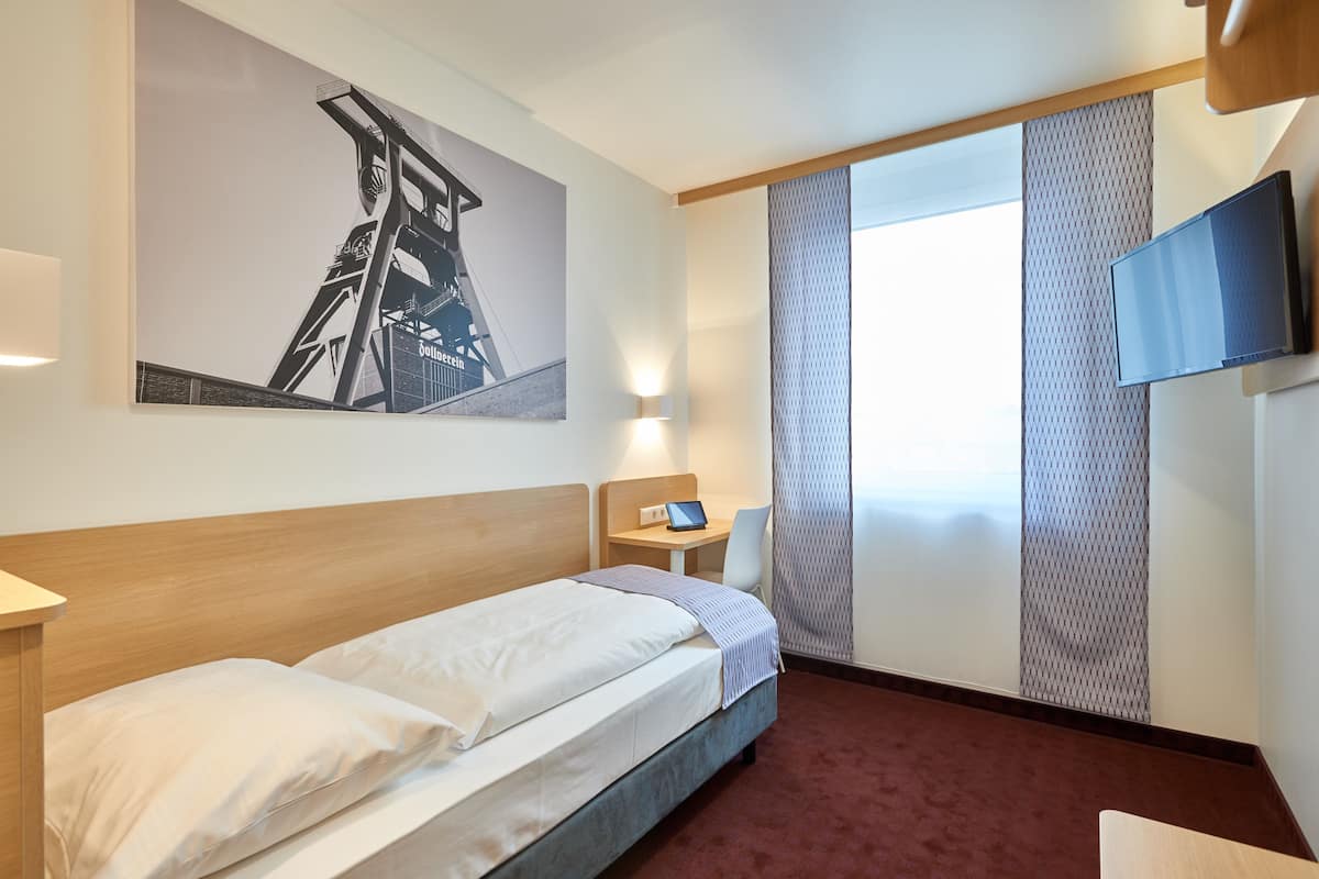 Une chambre d’hôtel avec un lit, un bureau, une chaise, une télévision et une grande fenêtre avec des rideaux.