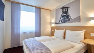 Una habitación de hotel con una cama grande, cabecera, mesita de noche, ventana con cortinas y un cuadro en la pared.