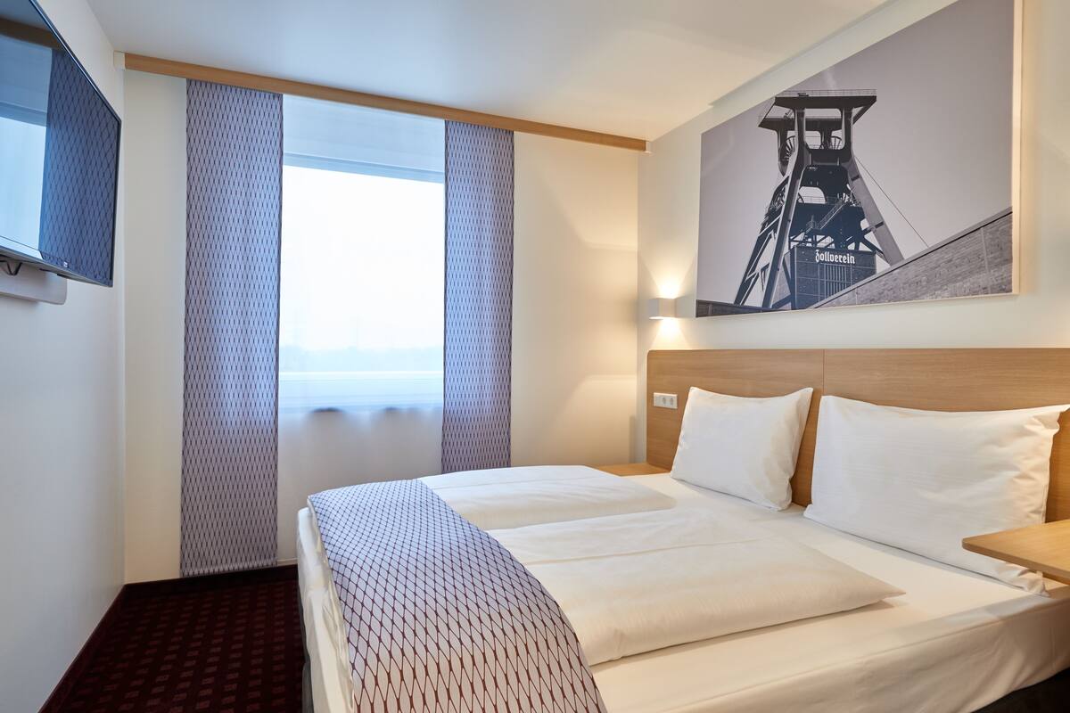 Une chambre d’hôtel avec un grand lit, un téléviseur fixé au mur et une fenêtre avec des rideaux.