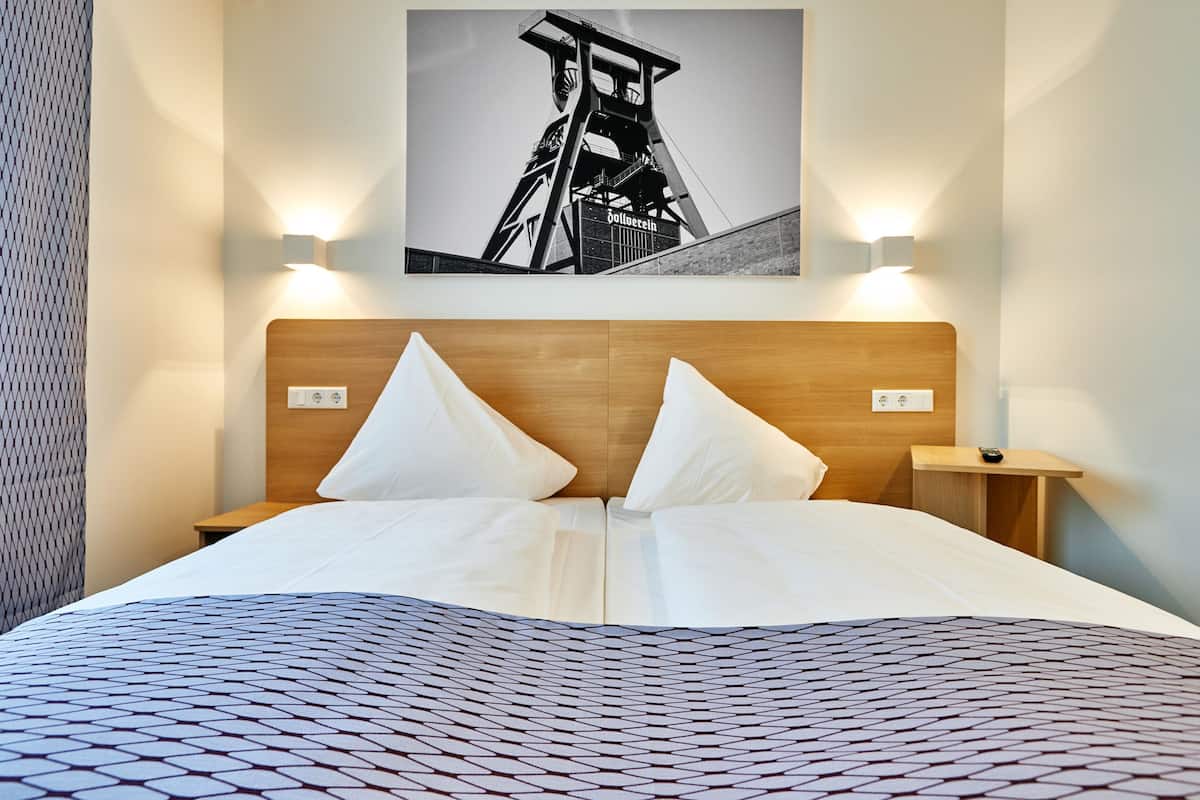 Une chambre d’hôtel avec un lit, deux oreillers, une tête de lit, une table de chevet, une lampe fixée au mur et un tableau encadré accroché au mur.