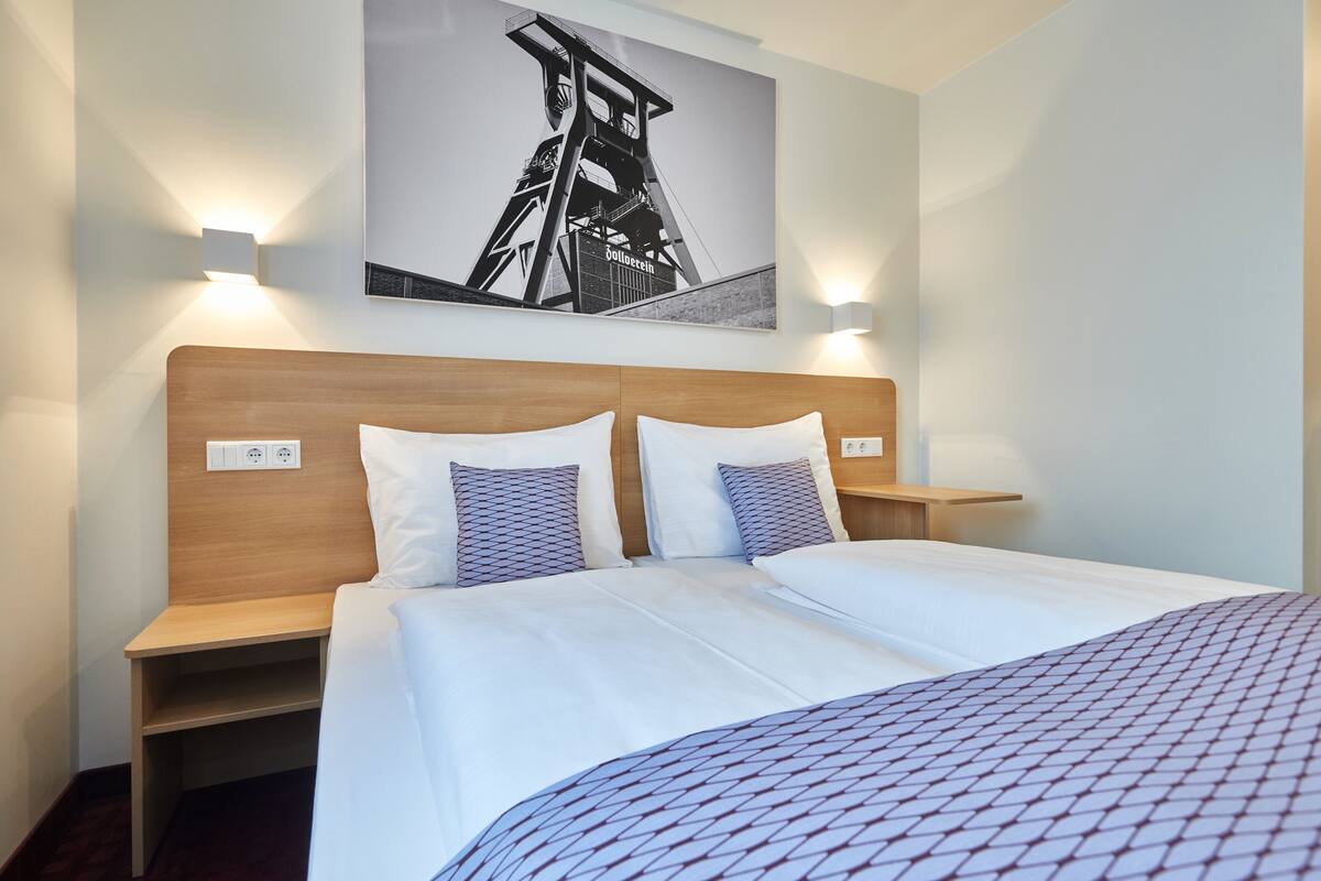 Une chambre d’hôtel avec une tête de lit en bois, deux lits avec du linge de lit blanc, des oreillers à motifs, des tables de chevet et un tableau accroché au mur.