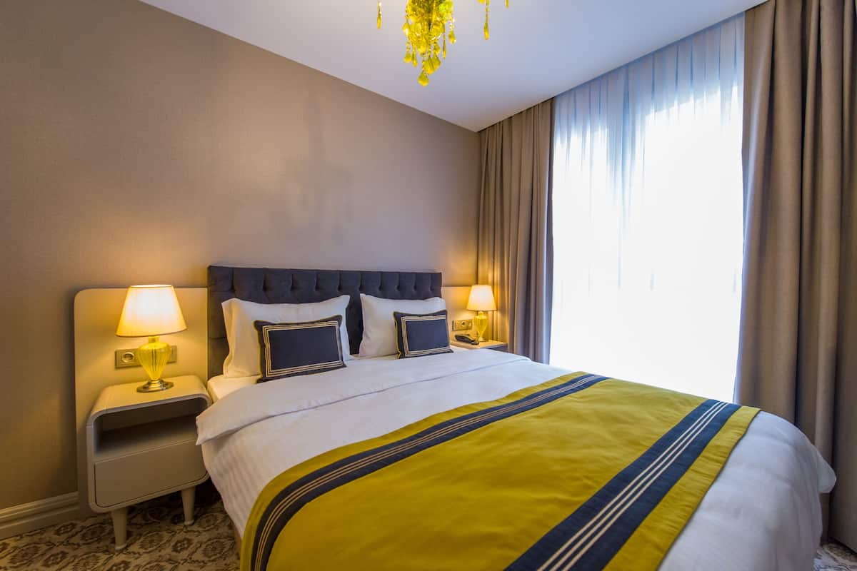Ein Hotelzimmer mit einem großen Bett, zwei Nachttischen mit Lampen, einer Kopfteil und Blick ins Freie durch ein Fenster mit Vorhängen.