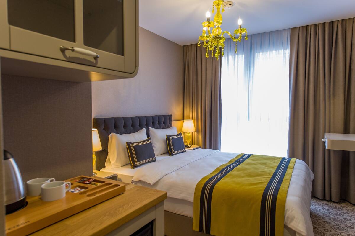 Ein Hotelzimmer mit einem Bett, Nachttischlampen, einem Holznachttisch mit Wasserkocher und Tassen, einer Kandelaber und einem großen Fenster mit Vorhängen.