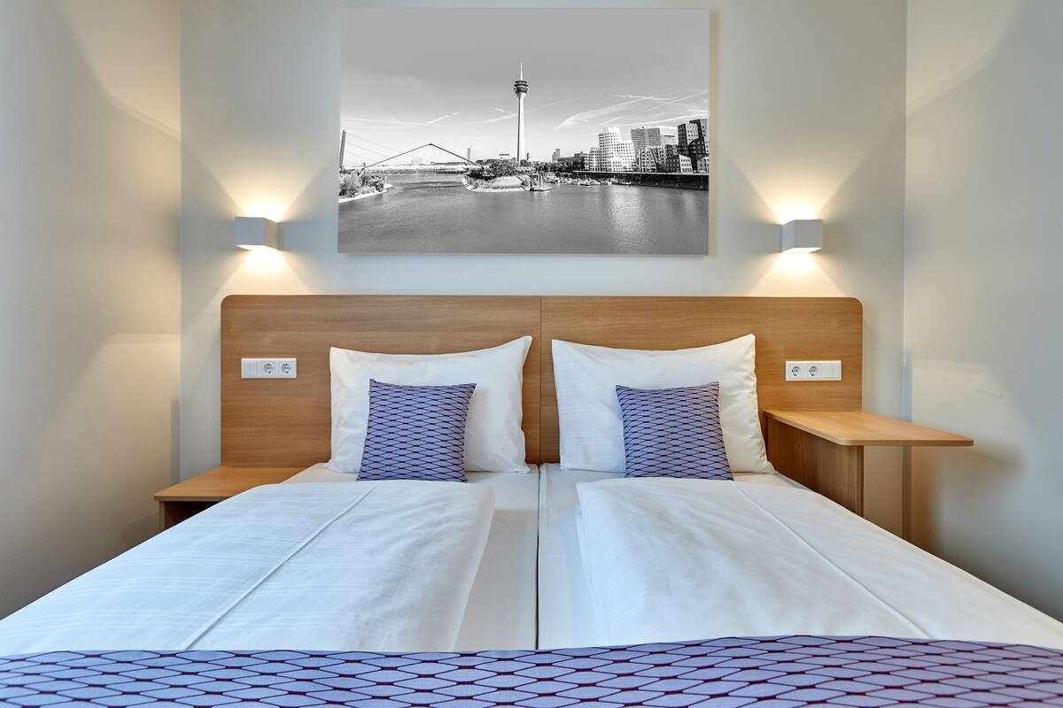 A double bed with white linens and patterned pillows, two bedside tables, and a cityscape photograph above.