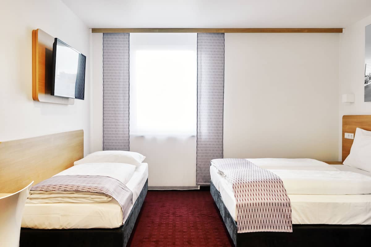 A hotel room with two beds, a TV mounted on the wall, and a large window with curtains.