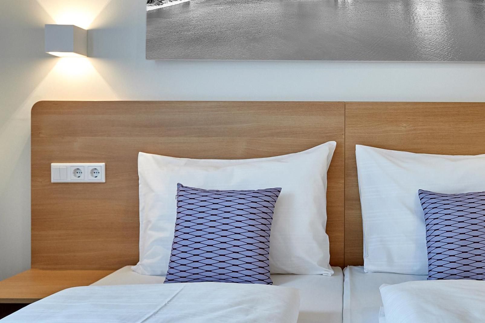 Une chambre d’hôtel avec une tête de lit en bois, deux oreillers blancs ornés d’un motif géométrique et un luminaire fixé au mur.